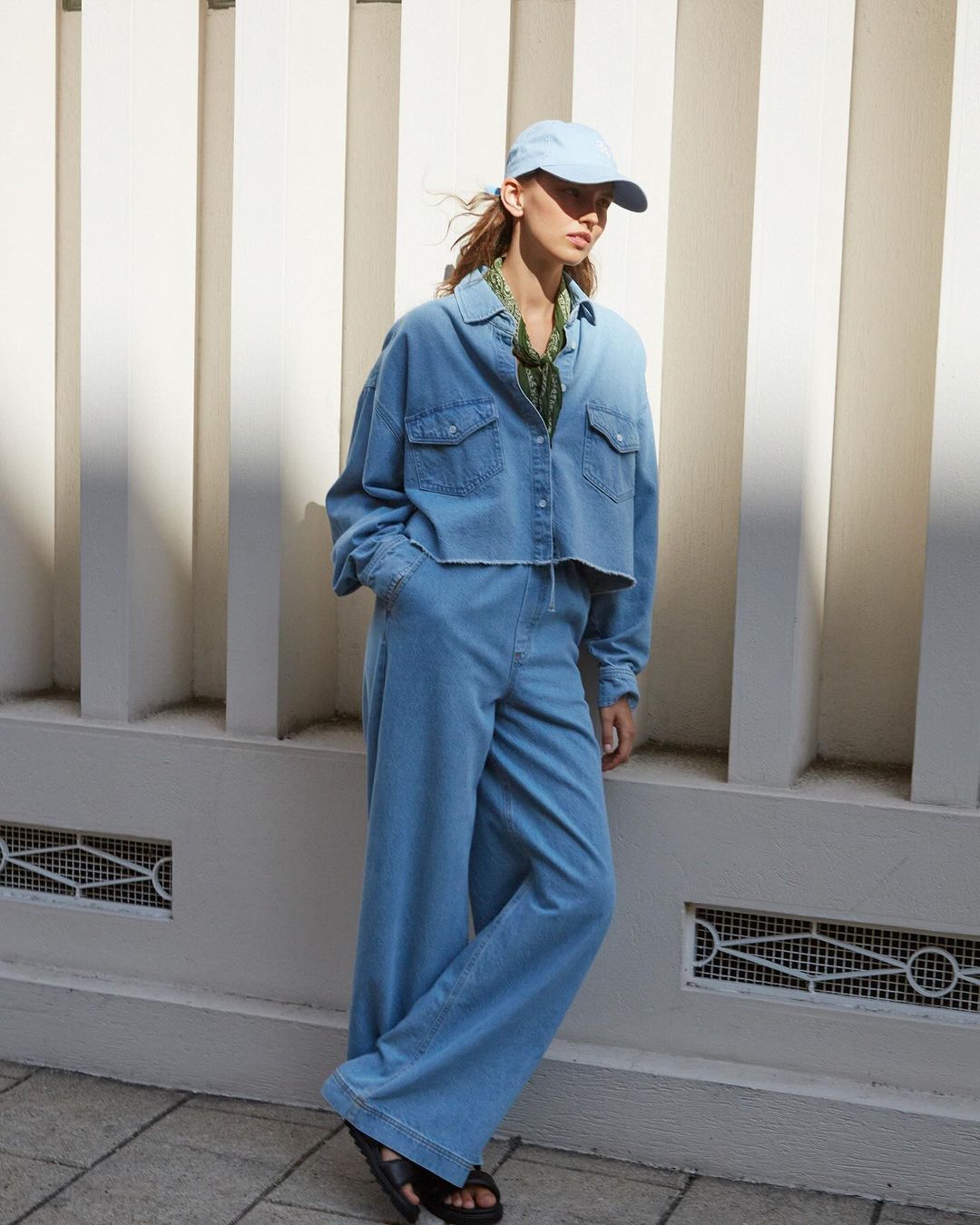 Model wearing a light blue denim set with matching cap posing against a pale architectural wall in Paris.