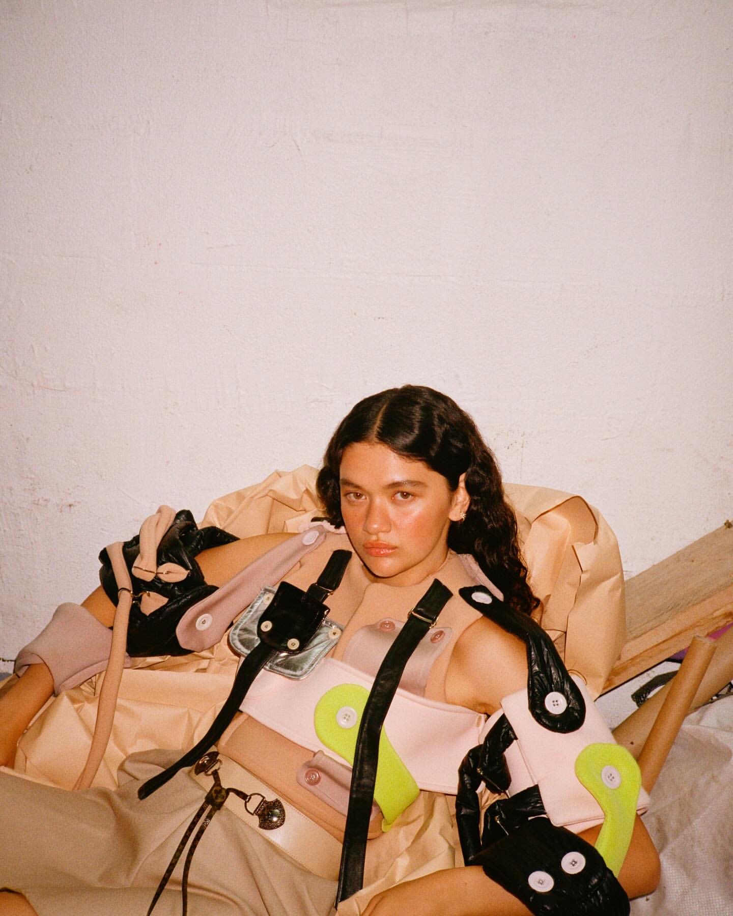 A young woman sits surrounded by deconstructed fashion items, wearing a unique, contemporary fashion ensemble with light and neutral tones.