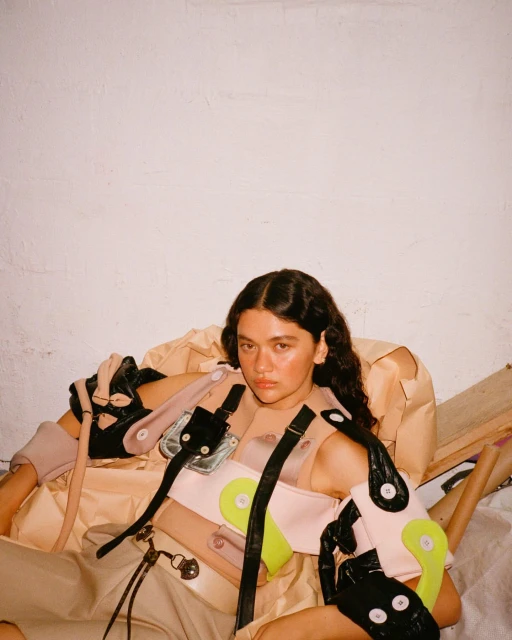 A young woman sits surrounded by deconstructed fashion items, wearing a unique, contemporary fashion ensemble with light and neutral tones.