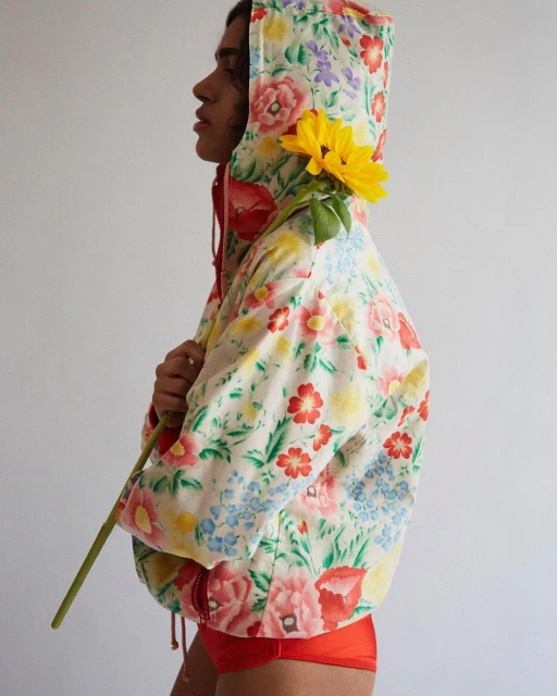 A person in a floral hoodie holds a sunflower against a white backdrop, showcasing a blend of fashion and nature-inspired design.