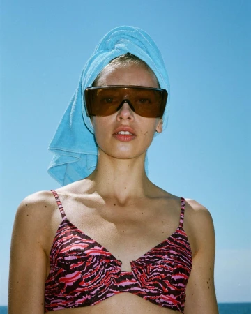Woman in a pink and black bikini top and oversized sunglasses, wearing a light blue towel on her head, with a clear blue sky background.