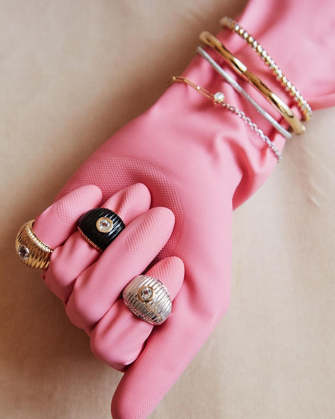 Hand wearing pink glove displaying multiple rings and bracelets in a close-up studio shot against a neutral background. Jewelry product shot.