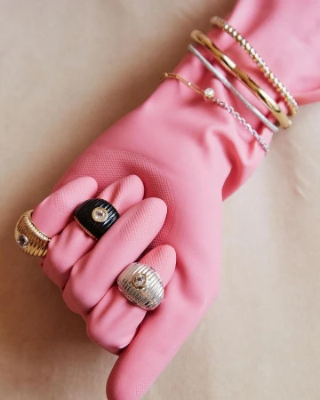 Hand wearing pink glove displaying multiple rings and bracelets in a close-up studio shot against a neutral background. Jewelry product shot.