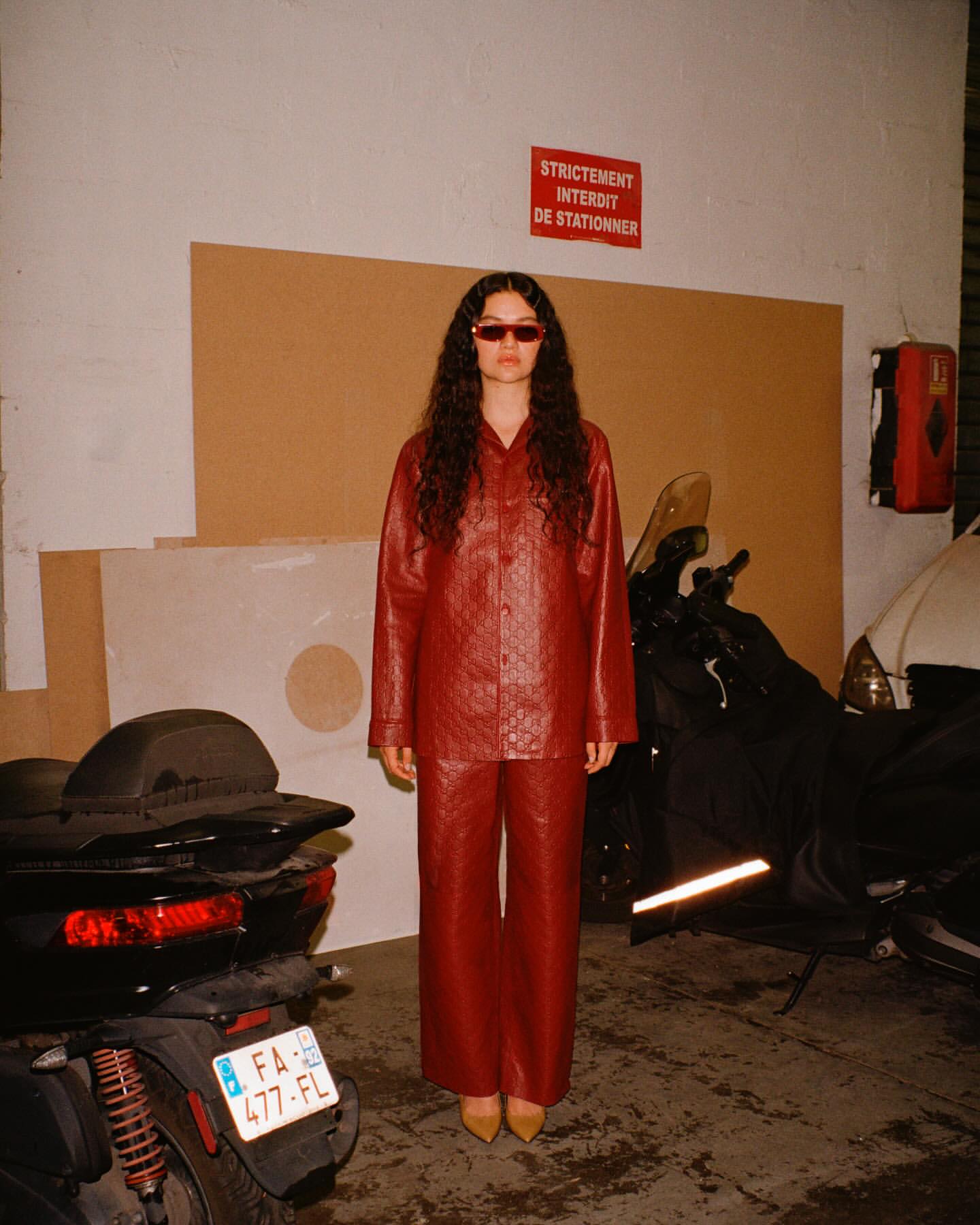 A young adult woman with sunglasses poses in a reddish-brown faux-leather outfit near a black scooter in an industrial setting.