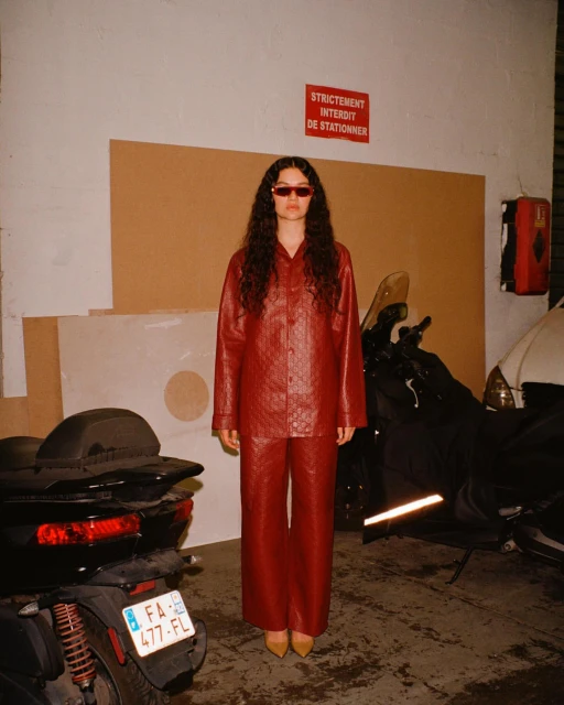 A young adult woman with sunglasses poses in a reddish-brown faux-leather outfit near a black scooter in an industrial setting.
