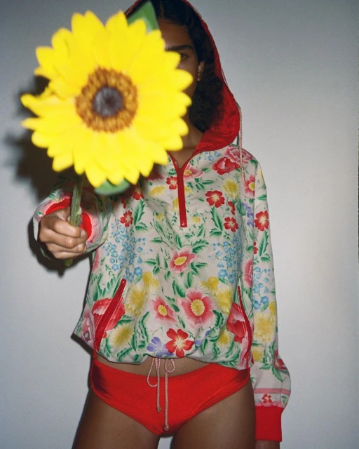 A person holds a large sunflower in front of their face, wearing a floral hoodie and red bottoms against a neutral backdrop.