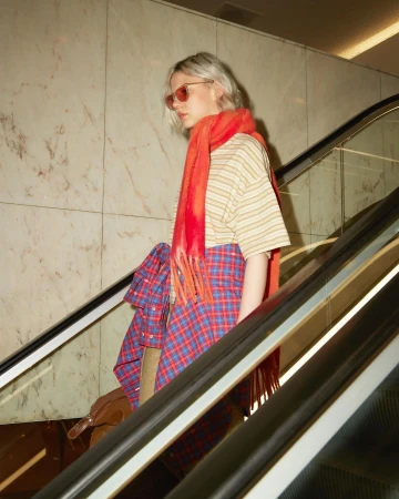 Stylish young woman on an escalator wearing an orange scarf, sunglasses, a striped shirt, and a plaid skirt, holding a brown bag.