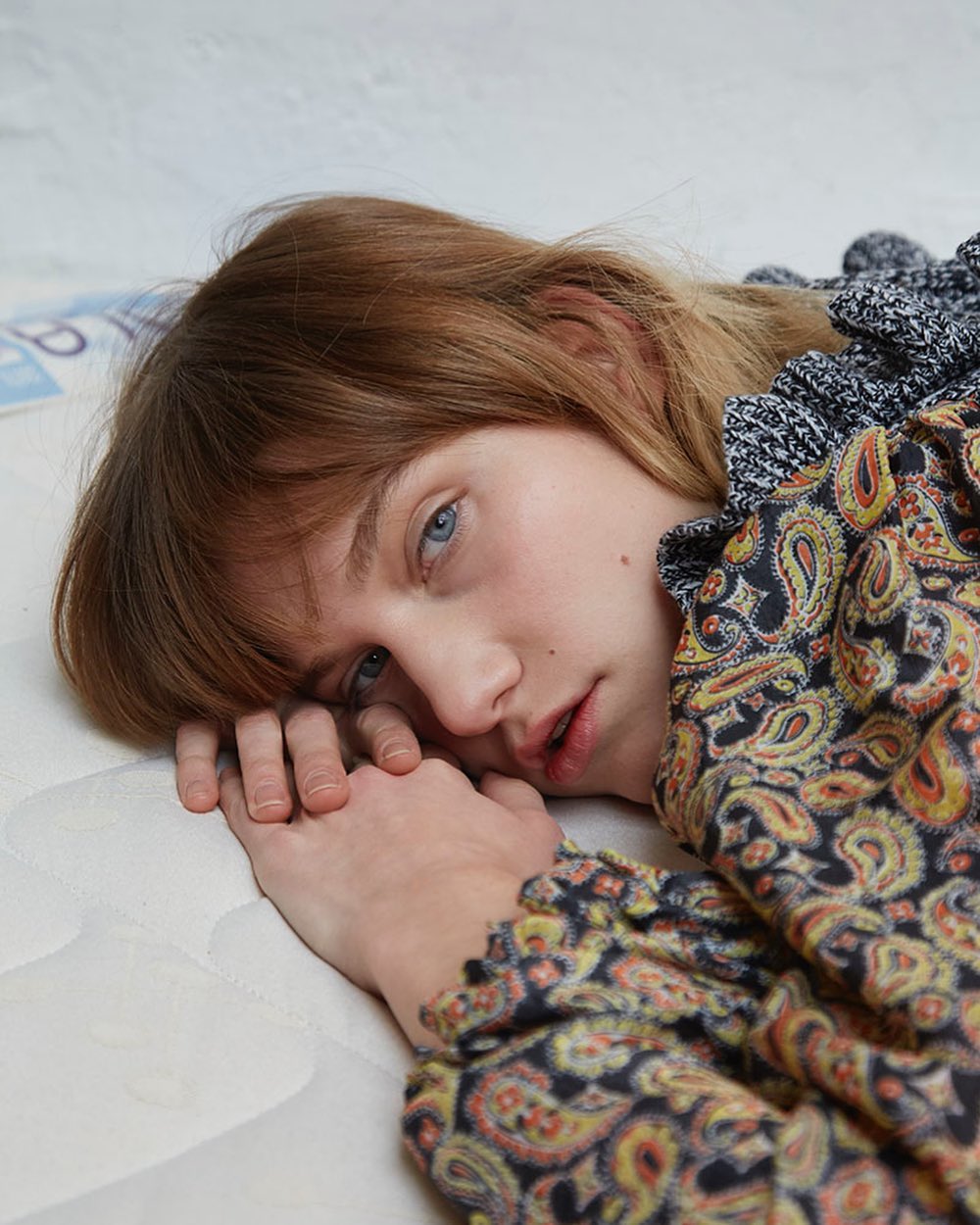 Close-up portrait of a young adult woman with reddish-brown hair, lying on a mattress, wearing a paisley print top with ruffled collar.