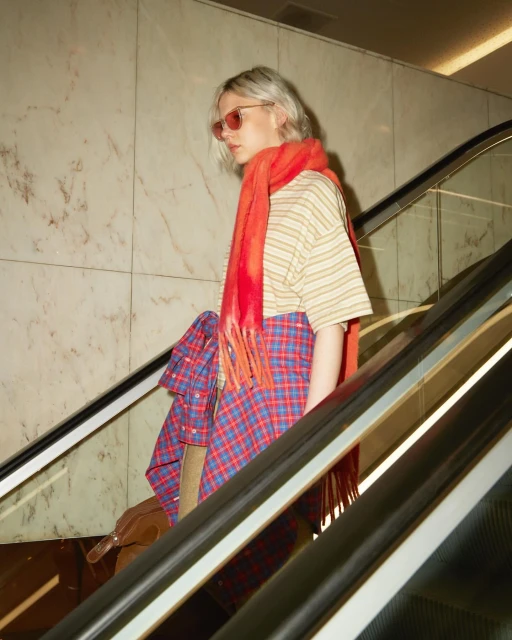 A young adult female with blonde hair wears a striped shirt, red scarf, and plaid skirt while standing on an escalator indoors.
