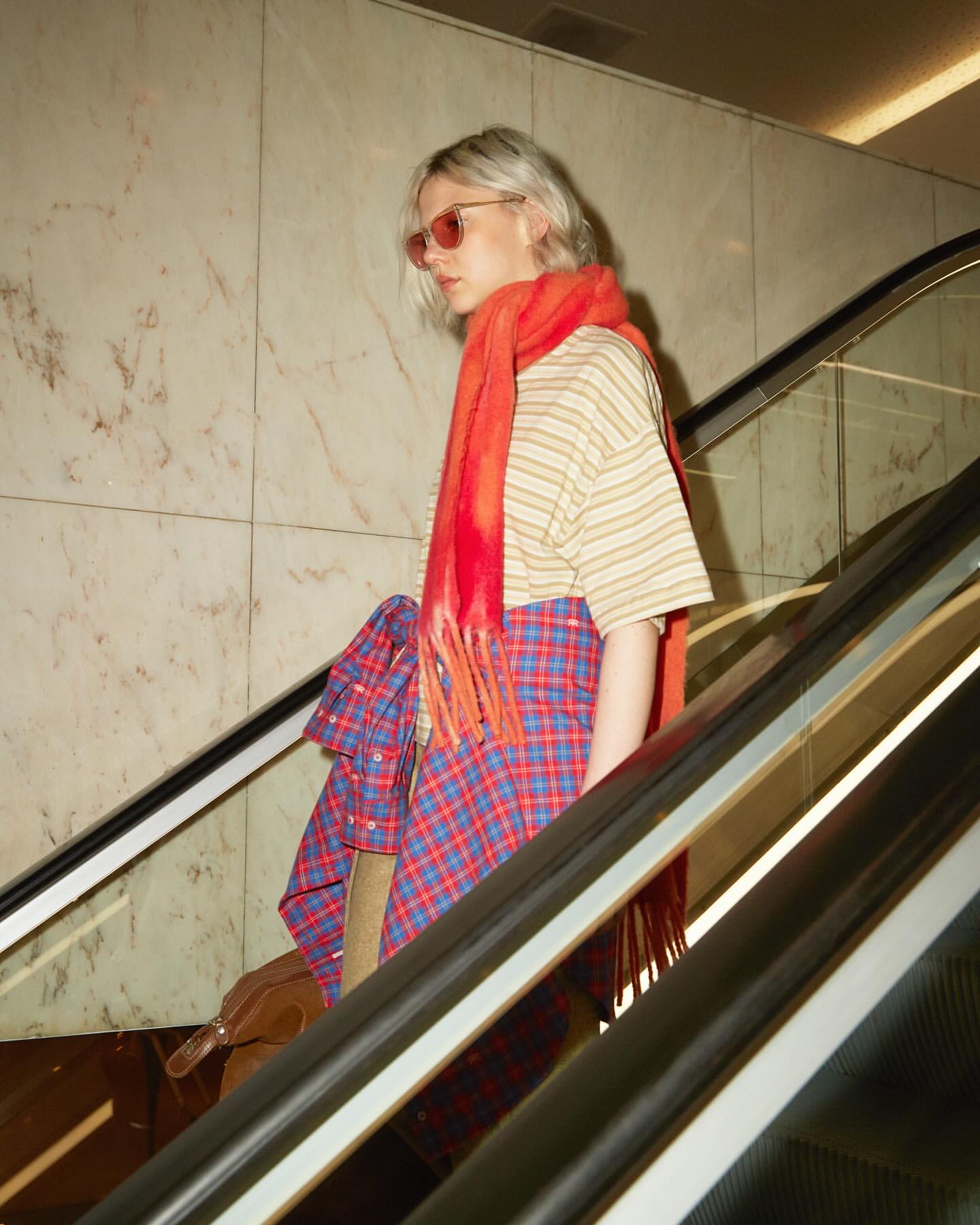 Woman on an escalator with a red scarf, wearing a striped shirt and plaid skirt, holding a brown bag, in an indoor environment.