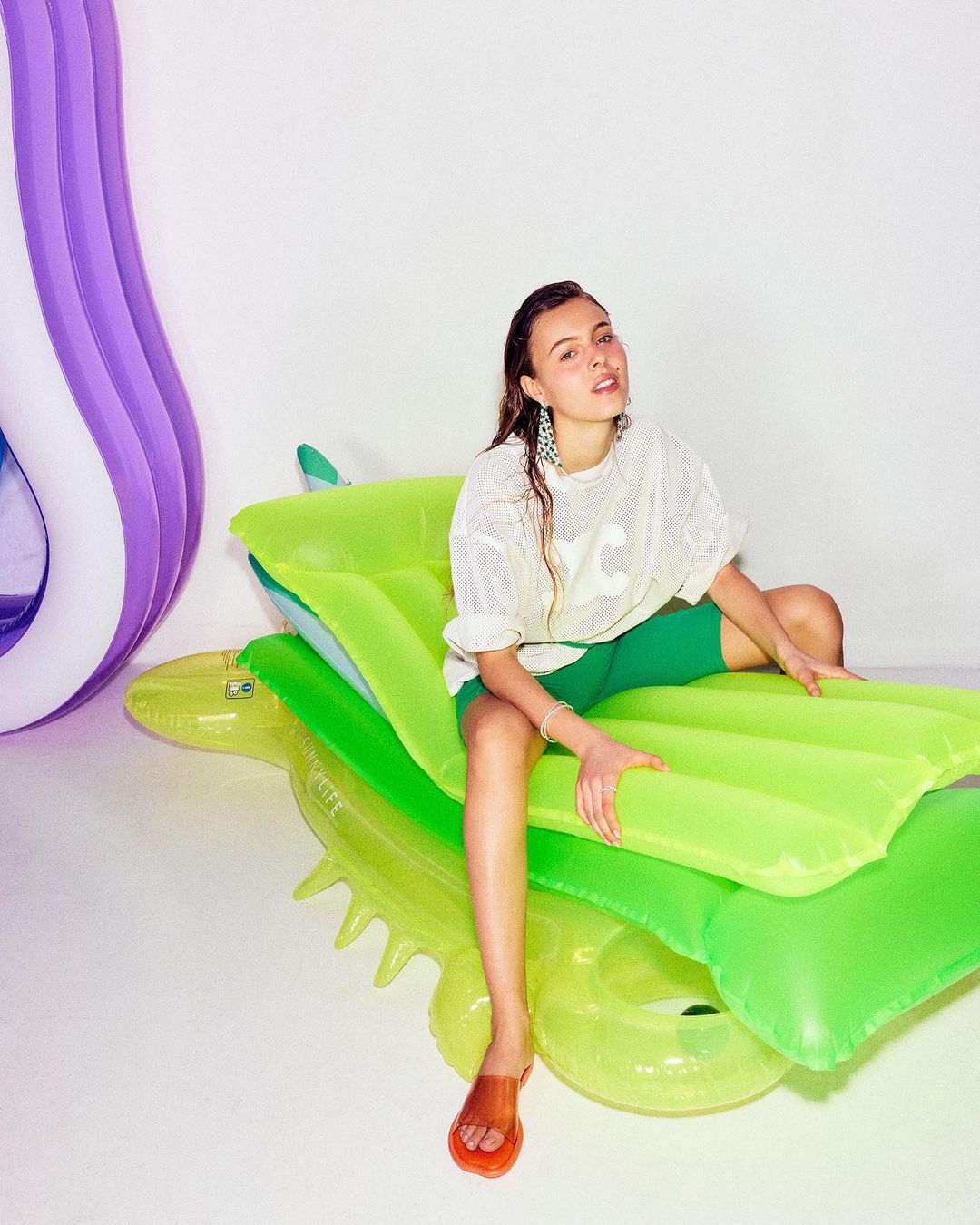 Young woman poses on a green inflatable crocodile, wearing a white t-shirt and green shorts, in a brightly lit studio.