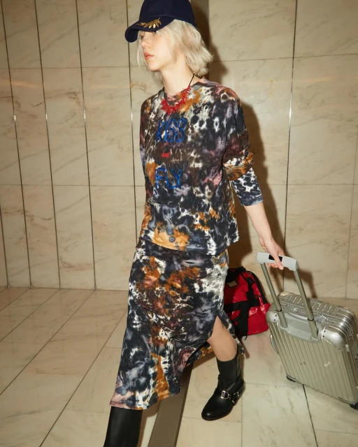 A fair-skinned young adult woman in patterned top and skirt pulls a silver carry-on suitcase across a tiled indoor floor.