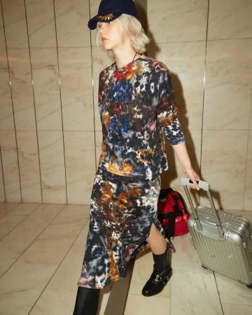 A fair-skinned young adult woman in patterned top and skirt pulls a silver carry-on suitcase across a tiled indoor floor.
