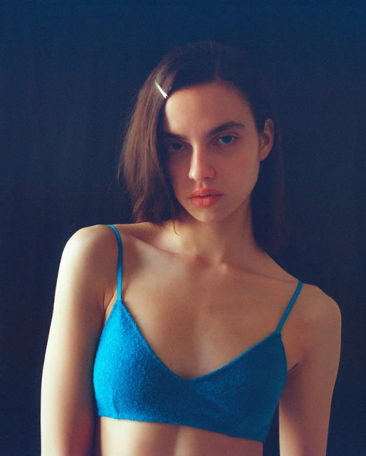 Portrait of a young woman with long brunette hair wearing a blue knitted bralette against a dark background in a studio setting.