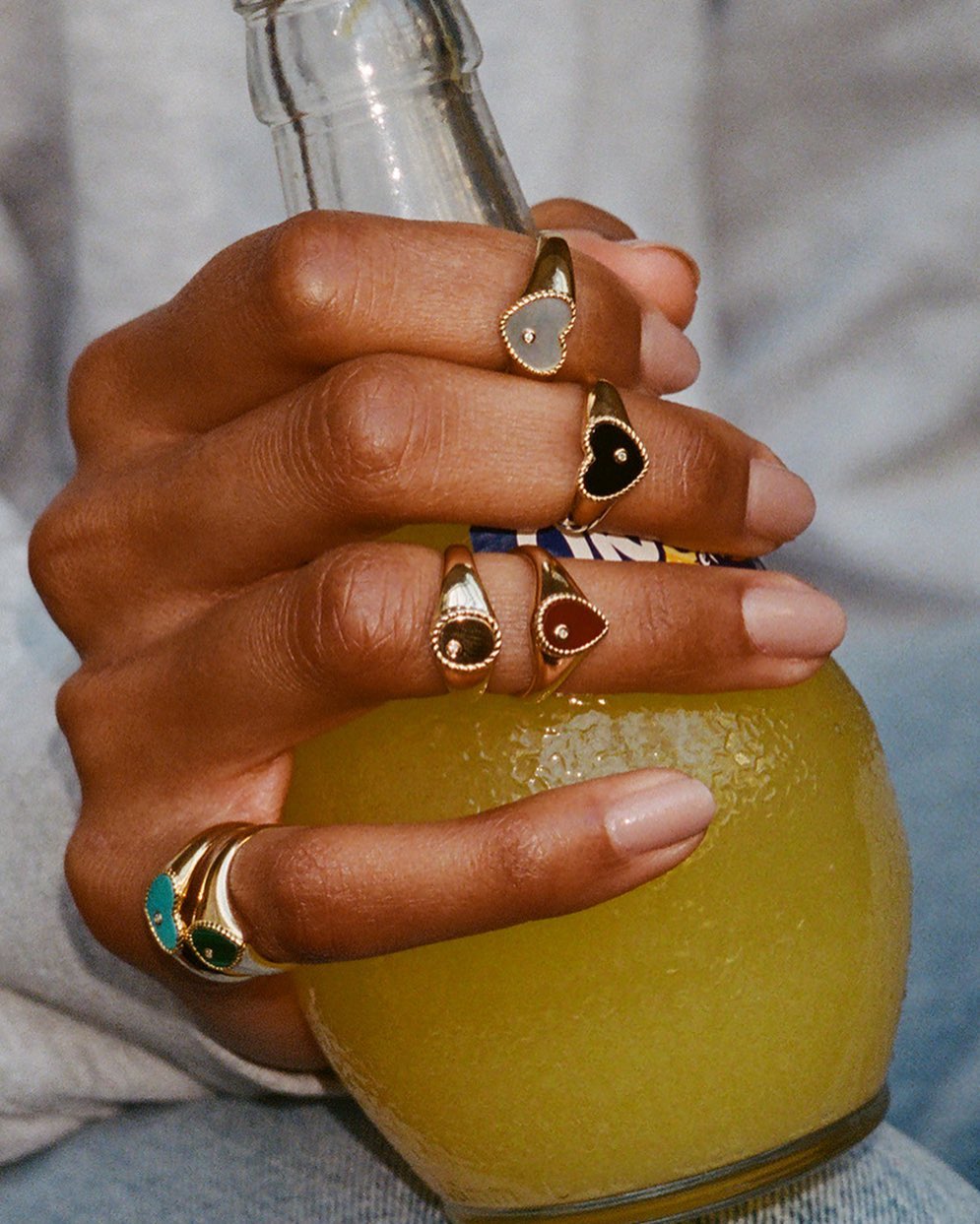Hand holding a soda bottle with multiple signet rings featuring heart and oval shapes on fingers, gold bands, and colored stones.