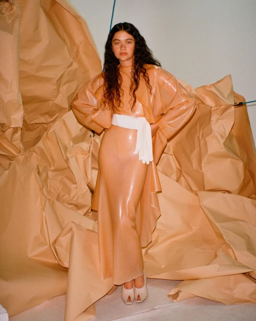 Young woman poses in a peach latex dress with a white belt, with crumpled brown paper in the background. Indoor fashion shot.