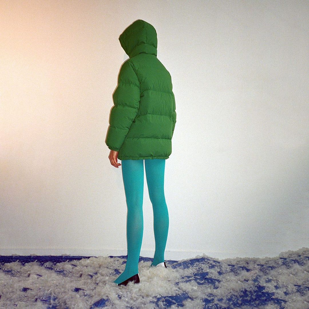 Woman in a green puffer jacket with blue tights stands on a bed of white feathers against a light wall, creating a striking visual.