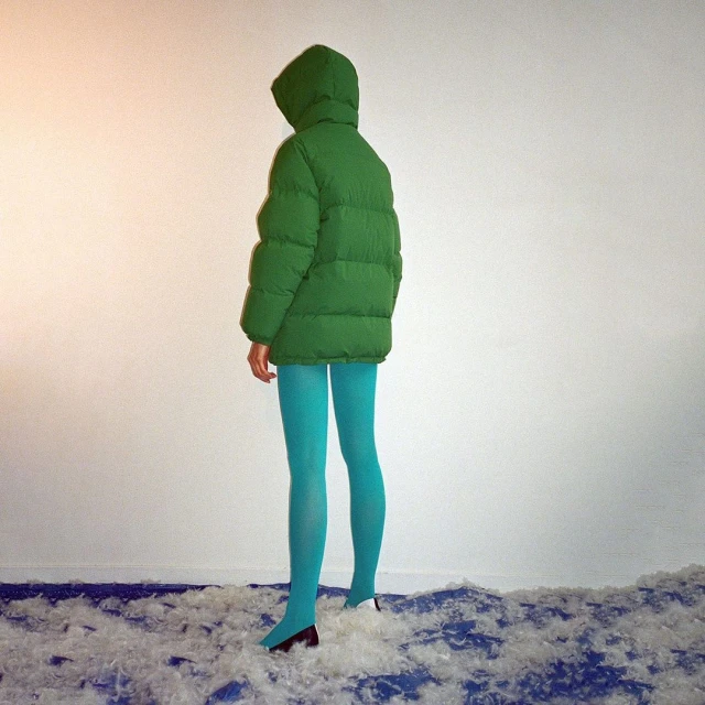 Woman in a green puffer jacket with blue tights stands on a bed of white feathers against a light wall, creating a striking visual.
