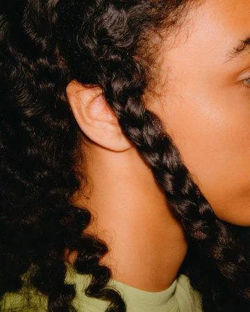 Close-up of a woman's face in soft lighting, featuring her dark curly hair styled with a single braid, and light green shirt.