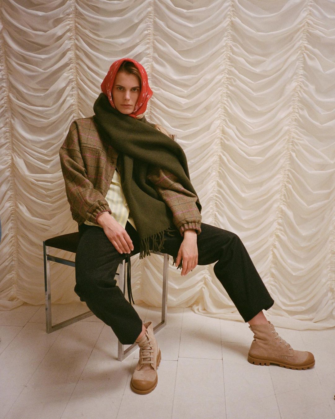 A person sits on a metal chair, wearing a brown tweed jacket, black pants, and tan boots, with a red headscarf and green scarf draped around their shoulders.