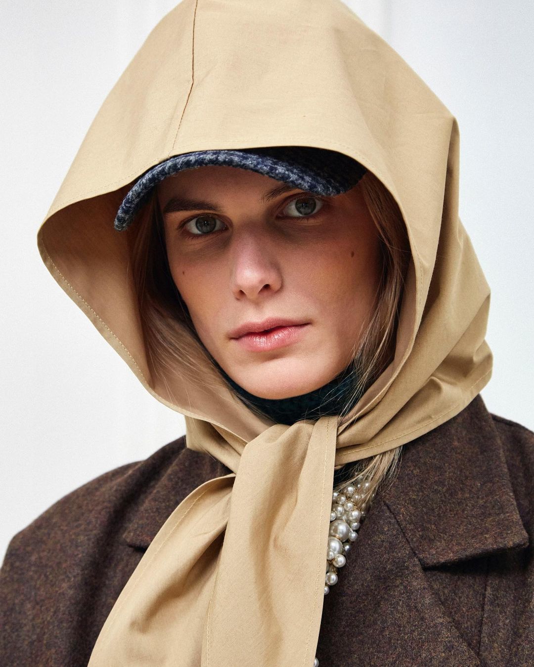 Fashionable adult female wearing a tan scarf and hat, brown jacket, and pearls in a studio shot against a plain white backdrop.