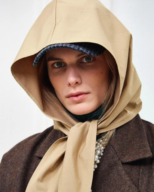 Fashionable adult female wearing a tan scarf and hat, brown jacket, and pearls in a studio shot against a plain white backdrop.
