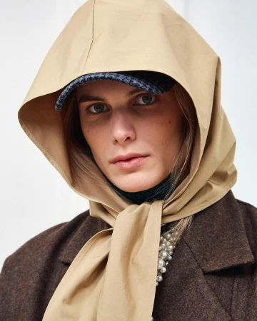 Fashionable adult female wearing a tan scarf and hat, brown jacket, and pearls in a studio shot against a plain white backdrop.