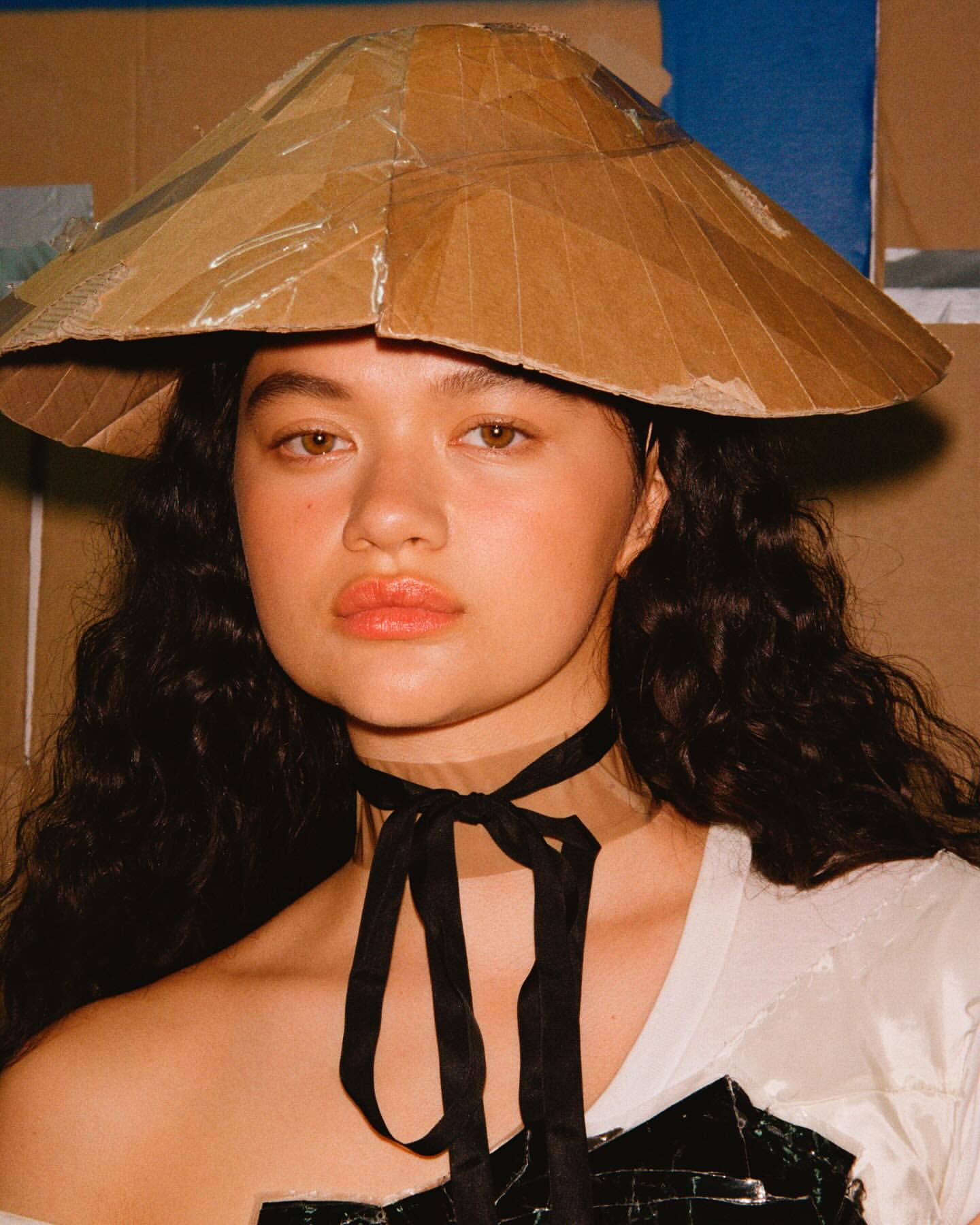 A young adult female model in a close-up shot wears an artistic cardboard hat and a black ribbon choker in a studio environment.