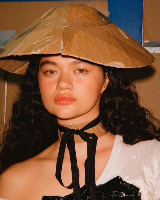 A young adult female model in a close-up shot wears an artistic cardboard hat and a black ribbon choker in a studio environment.