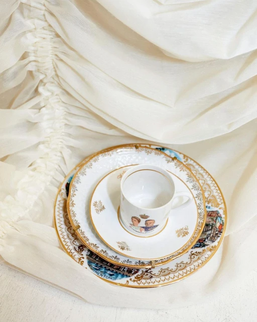 Elegant still life composition featuring a white porcelain cup and ornate plates with golden details, set against a flowing white cloth.