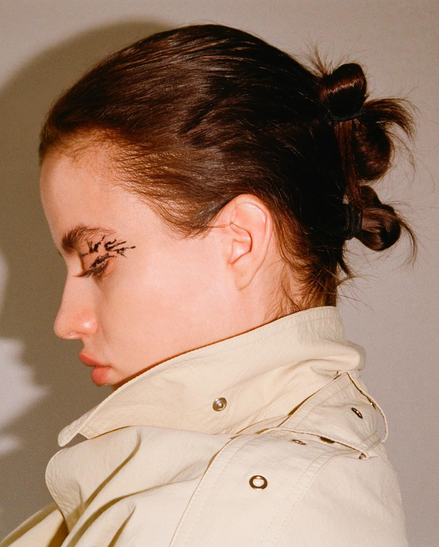 Close-up portrait of a woman with dark hair styled in small buns, wearing a beige jacket and black eyeliner on her eyelids.