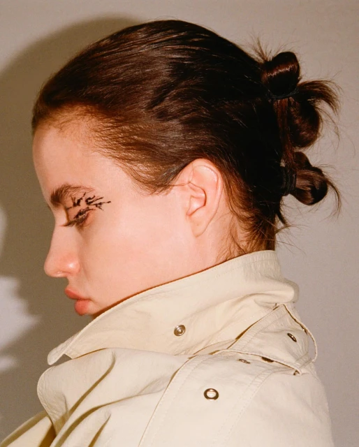 Close-up portrait of a woman with dark hair styled in small buns, wearing a beige jacket and black eyeliner on her eyelids.