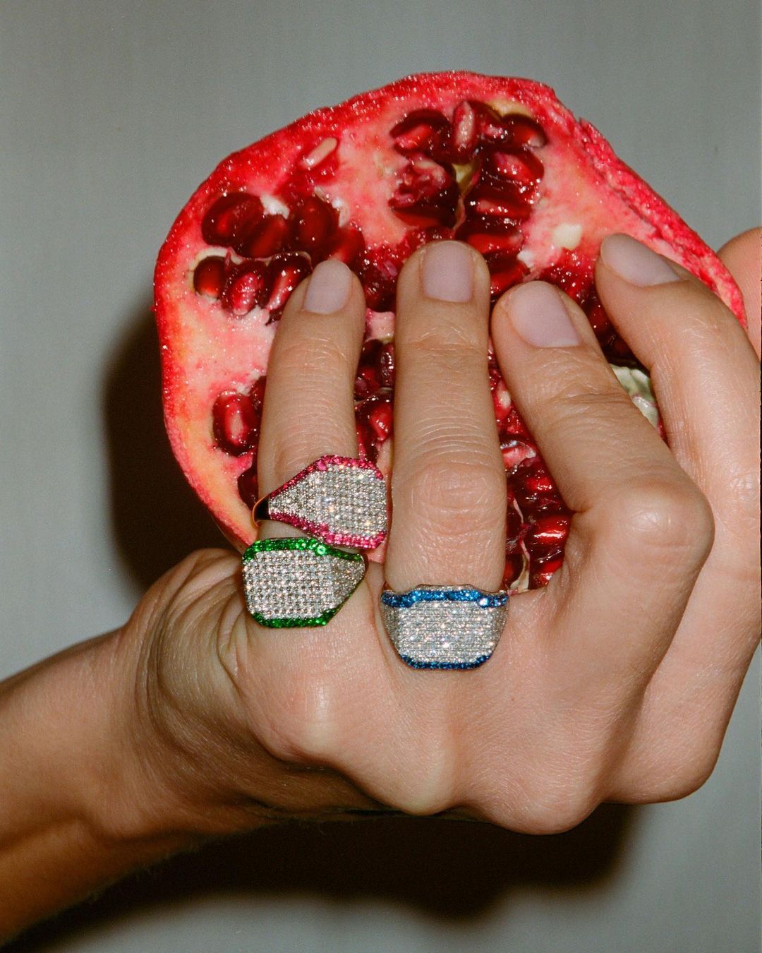 Close-up of a hand holding a halved pomegranate, wearing three diamond signet rings with ruby, emerald, and sapphire borders.