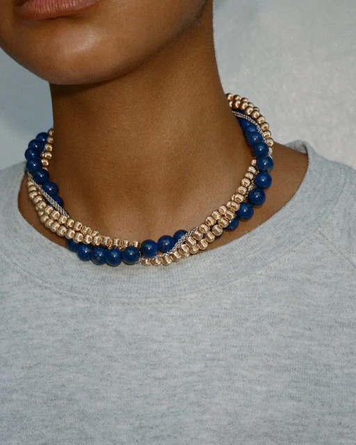 Person wearing a layered necklace with blue and gold beads. Close-up studio shot of fashion jewelry and a grey top.
