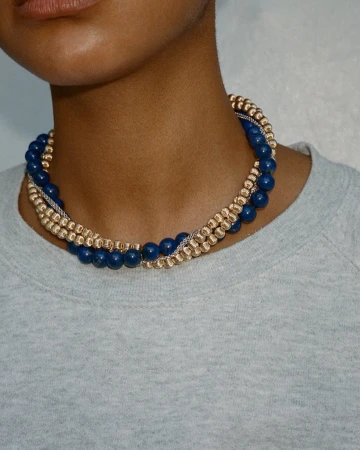 Person wearing a layered necklace with blue and gold beads. Close-up studio shot of fashion jewelry and a grey top.