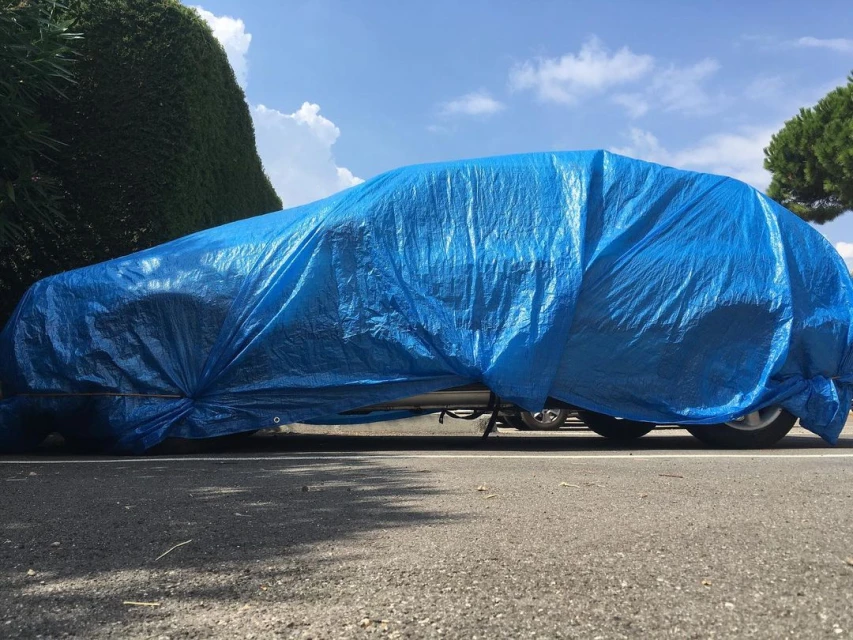 Car covered with a blue tarp parked outdoors on asphalt on a sunny day with green hedges and a blue, cloudy sky.