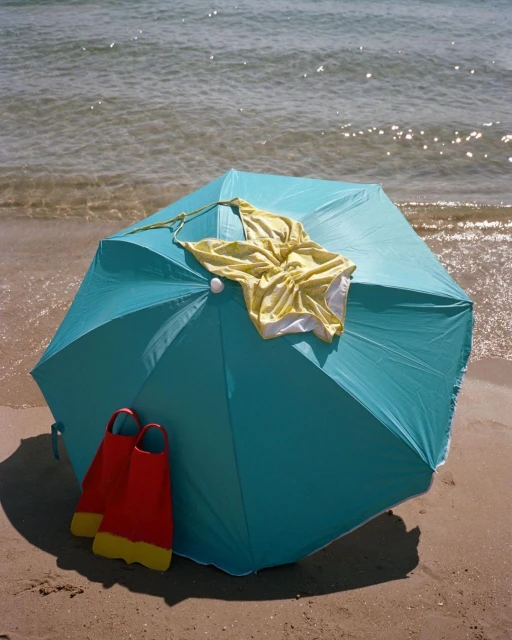 Aqua-blue beach umbrella with yellow swimsuit and red flippers on a sunny beach, with ocean waves softly breaking in the background.