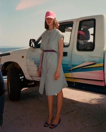 Woman in striped shirt dress and pink cap poses next to truck with painted design on sunny day. Fashion editorial photography.