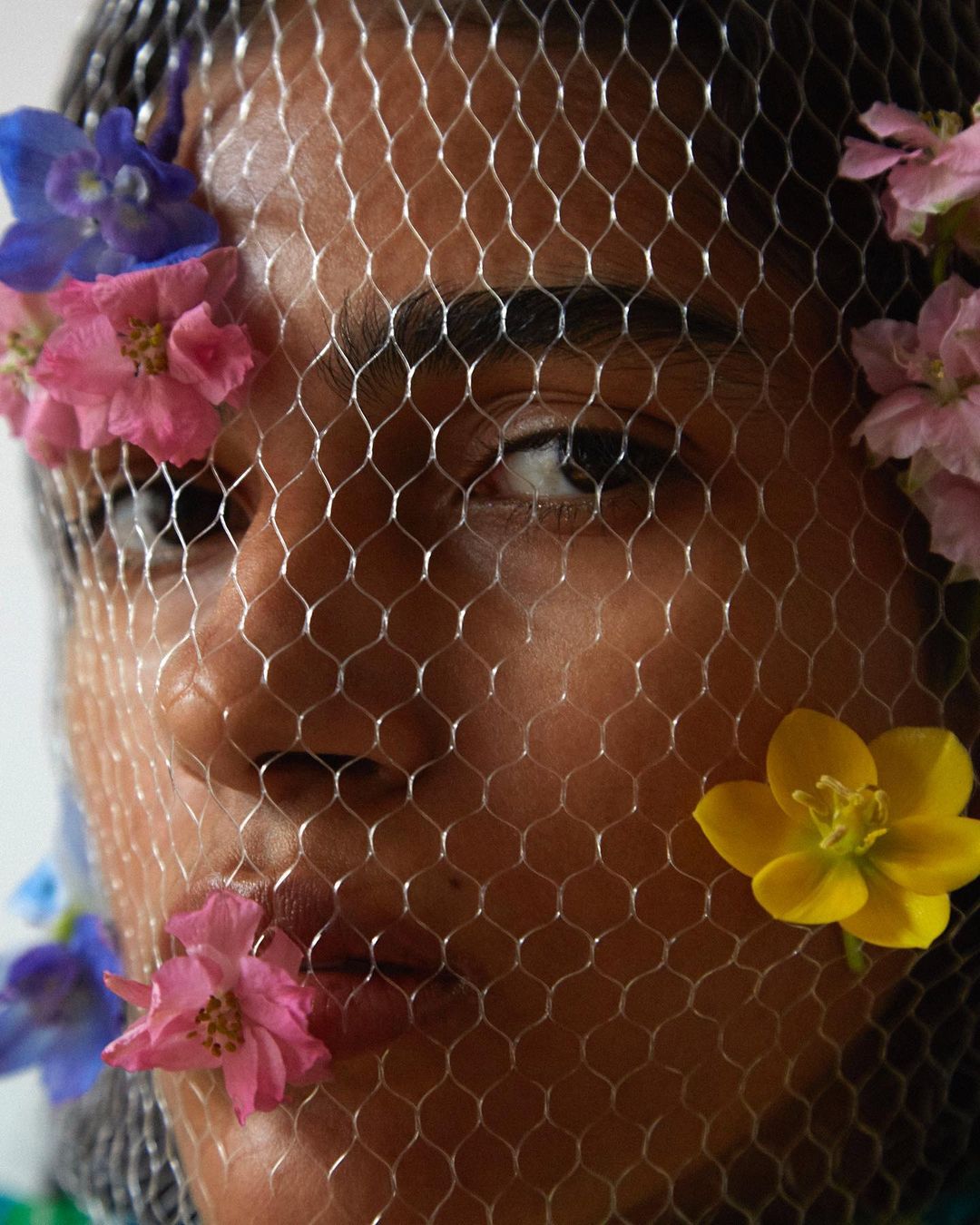 Close-up studio portrait of a dark-skinned woman wearing a silver net veil decorated with colourful blossoms and flower petals.