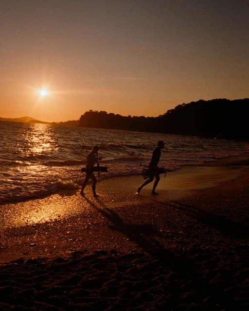 Two silhouetted figures carrying long objects walk along a beach at sunset, with the sun reflecting on the water and wet sand.
