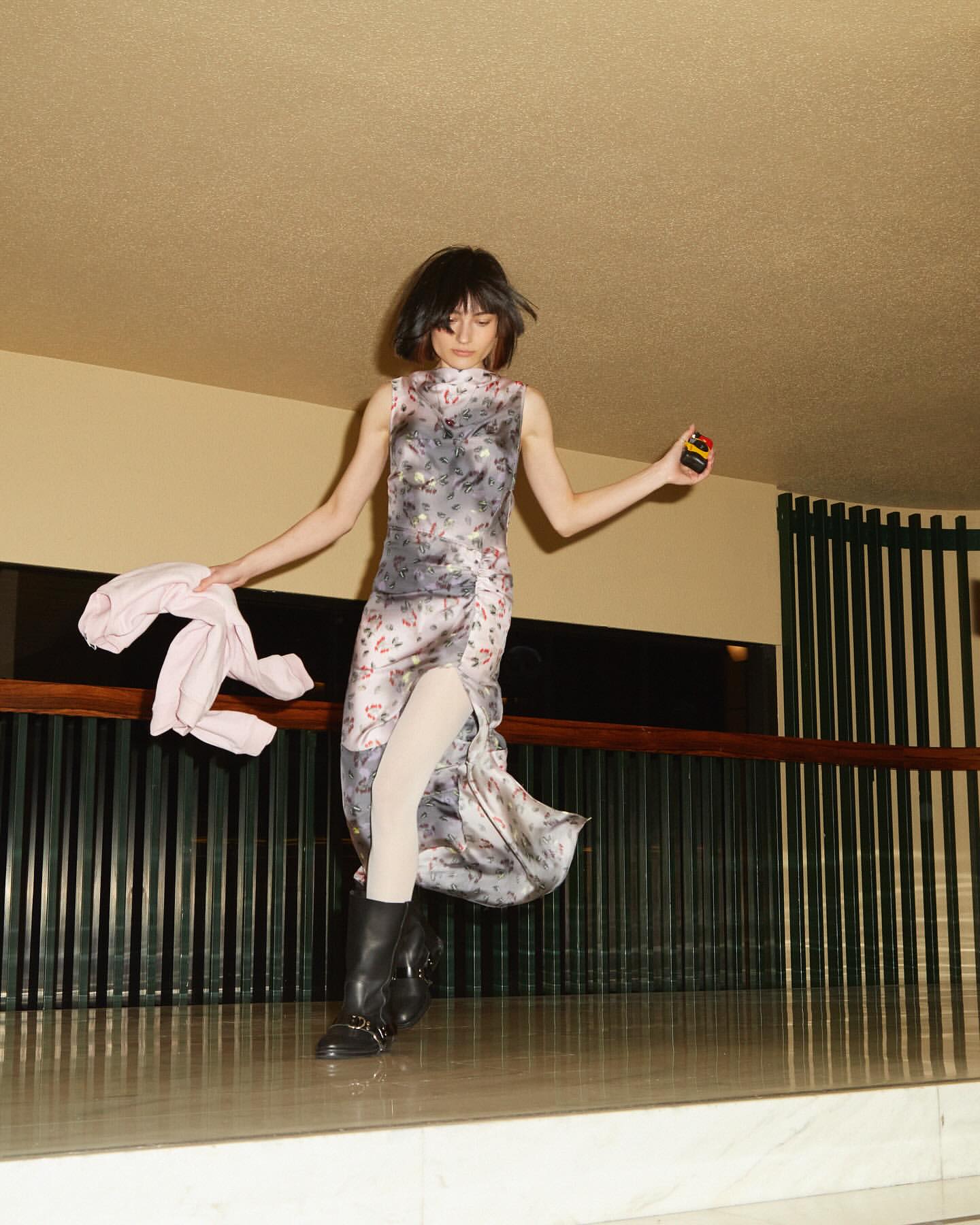 Woman in floral dress and boots poses indoors, holding a pink jacket and a small box, illuminated by warm, overhead lighting.