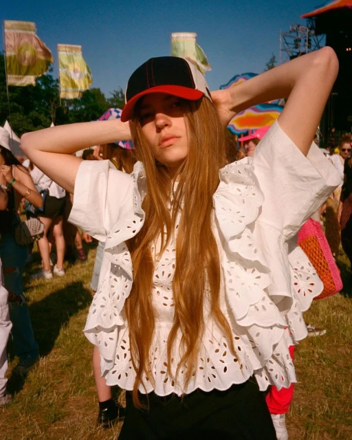 A woman with long brown hair in a white eyelet blouse and baseball cap stands in a grassy field at a daytime outdoor music festival.