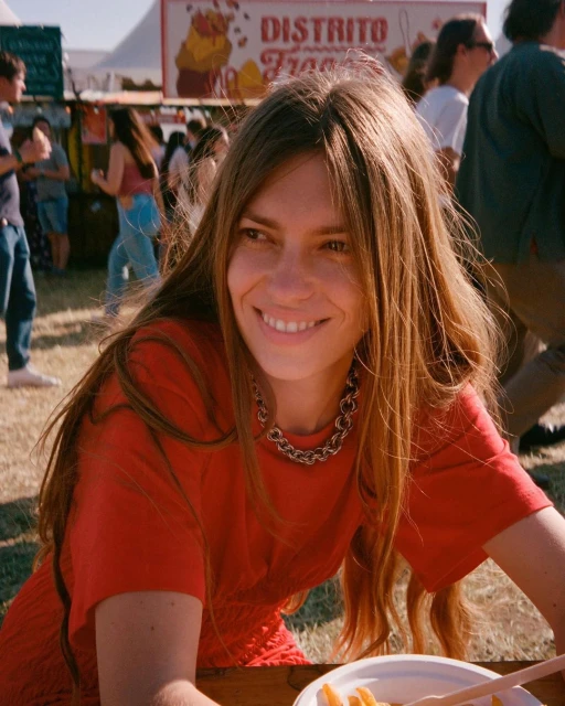 Smiling woman in a red dress sits at a table with food in an outdoor setting, with blurred people and a food stand in the background.