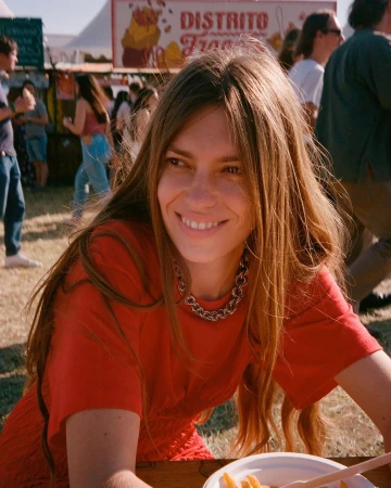 Smiling woman in a red dress sits at a table with food in an outdoor setting, with blurred people and a food stand in the background.
