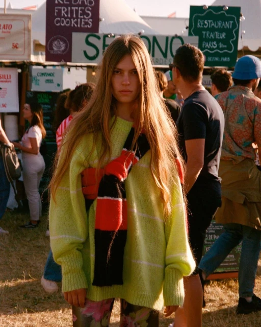 Teen with long hair wearing a lime green sweater and floral pants standing in front of food vendors and a crowd of people outdoors.