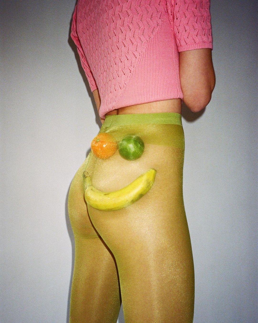 Model wears sheer yellow tights and pink top with fruit detail. A playful fashion image featuring banana, lime and mandarin.