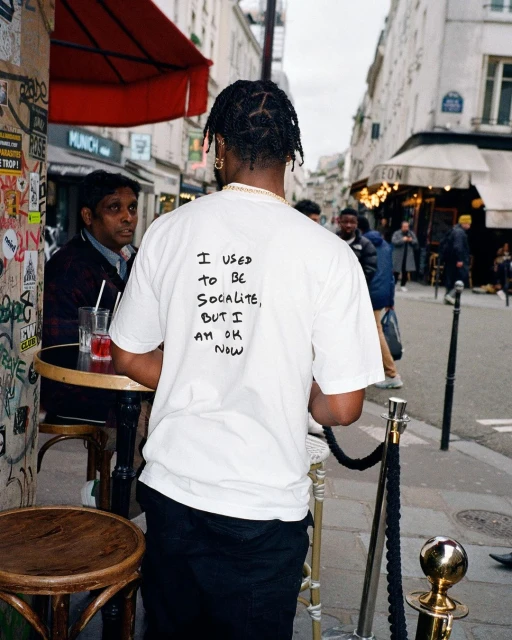 A man stands in a European city street, wearing a white t-shirt with handwritten text, near an outdoor cafe with people and details.