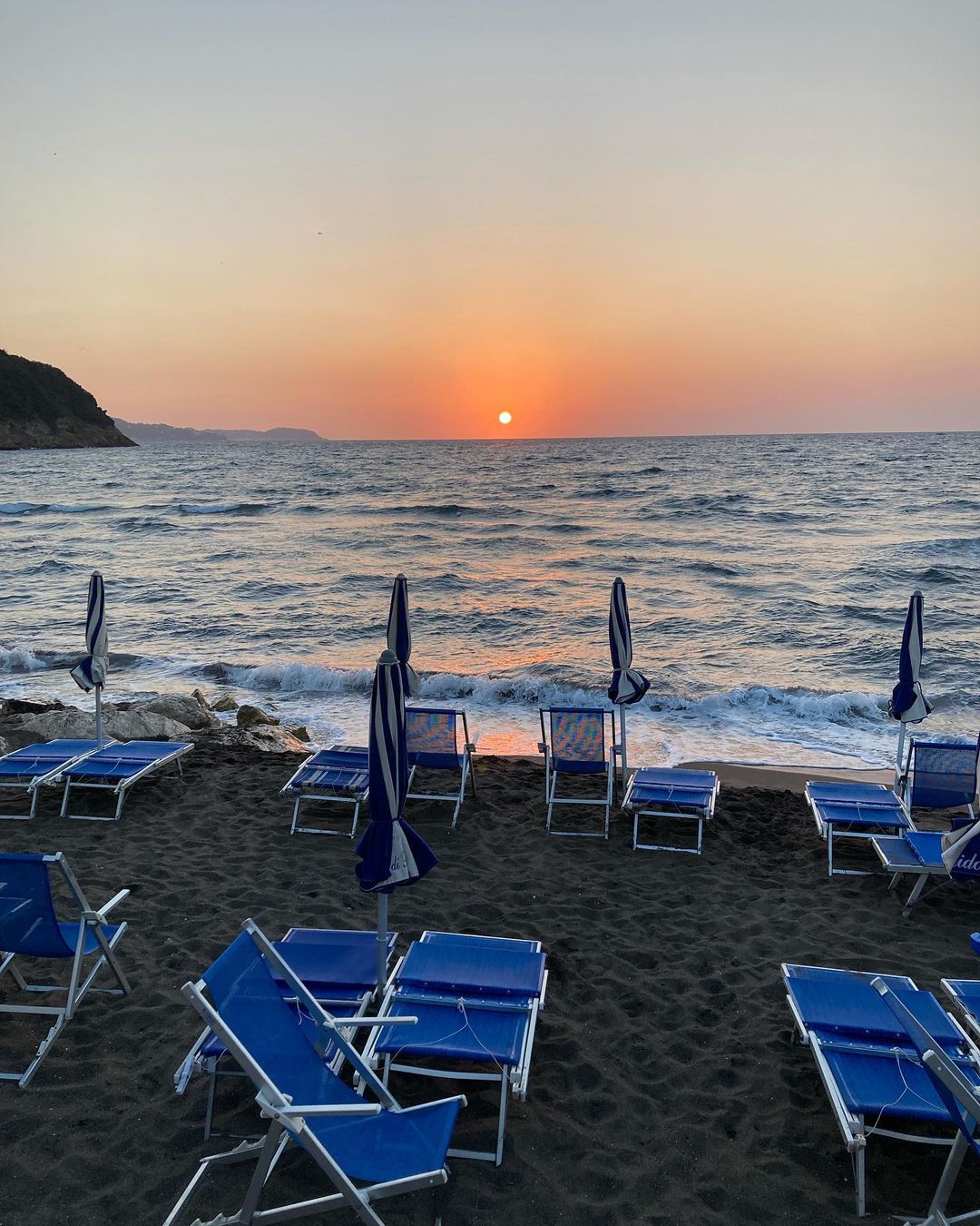 Sunset at a beach with multiple blue and white striped umbrellas and beach chairs. Waves are gently lapping at the shore.