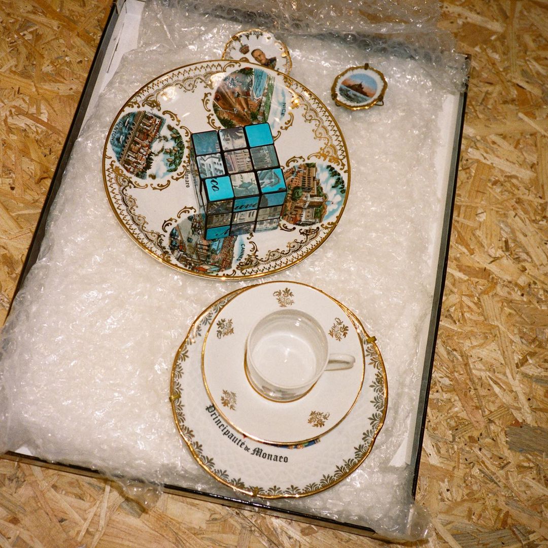 Rubik's cube resting on an ornate china plate alongside a teacup, saucer, and miniature plate, all encased in clear bubble wrap.