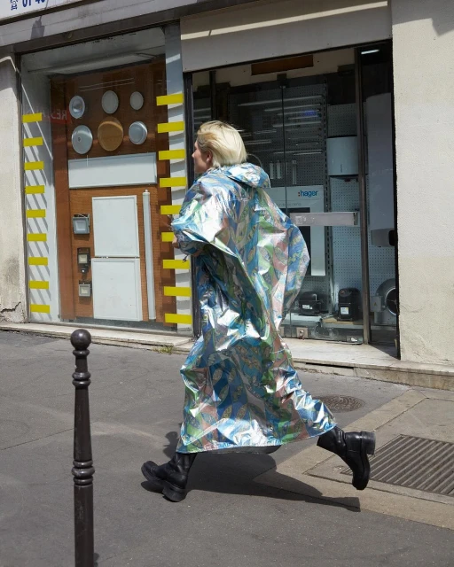 Side view of a slender adult female in a reflective silver hooded raincoat, walking on an urban street by a storefront.